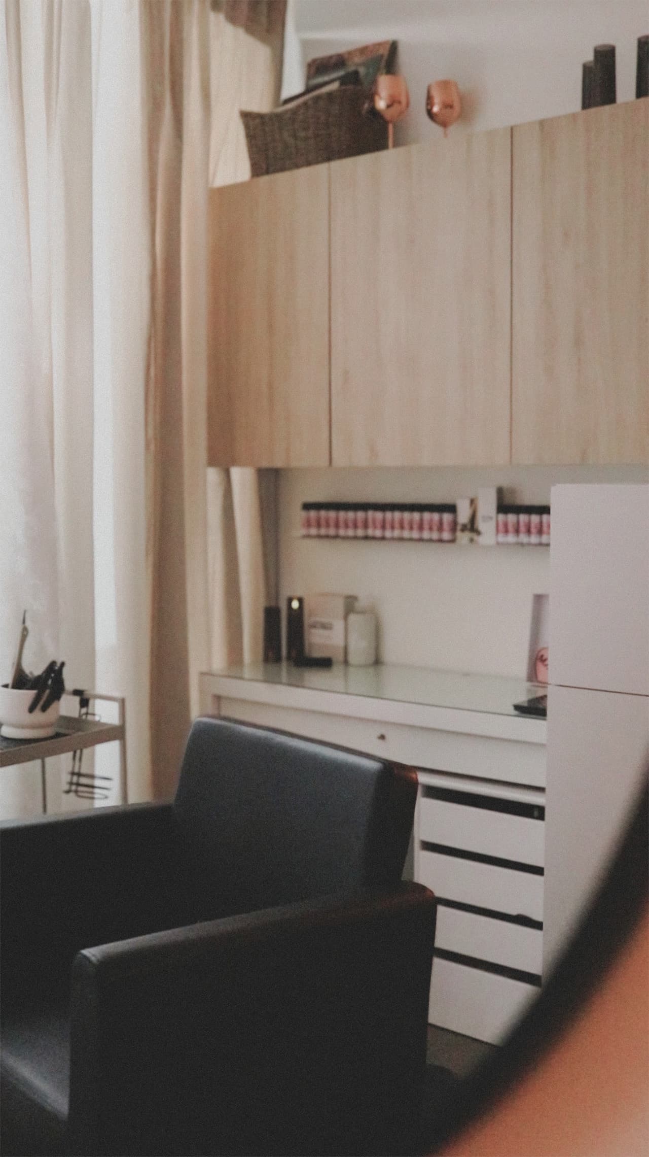 A black styling chair by sheer curtains in The Sanctuary, with wooden cabinets and a white vanity behind it