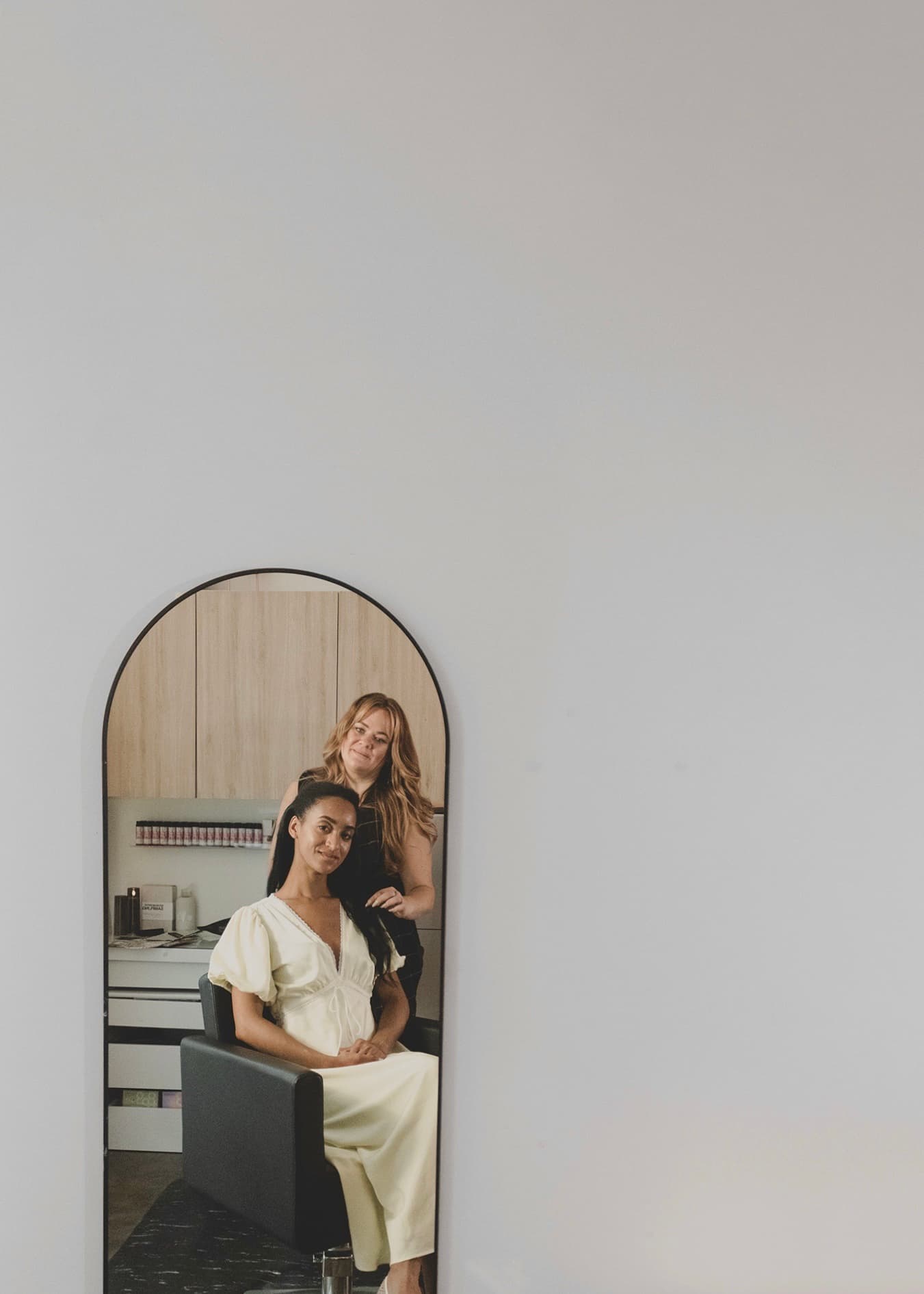 Reflected in an arched mirror, a guest in a cream dress sits in the styling chair as Steph stands behind her