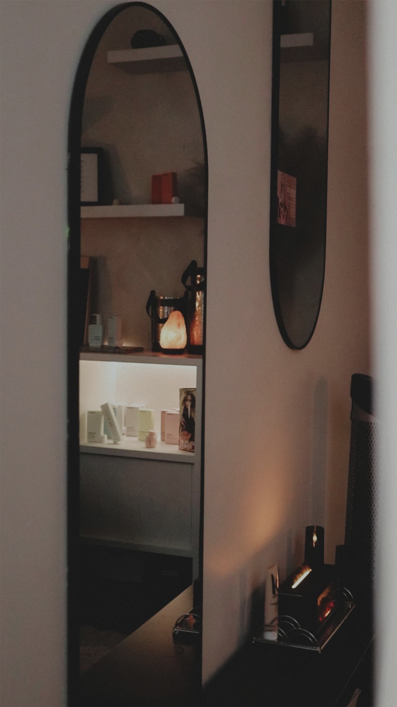 A tall arched mirror in The Sanctuary reflecting the lit shelves and salt lamp, in soft evening light