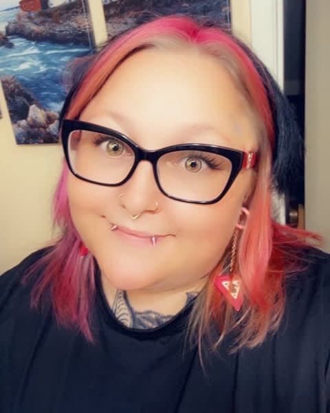 Dani Hauk, tattoo artist, with pink hair and black cat-eye glasses, smiling