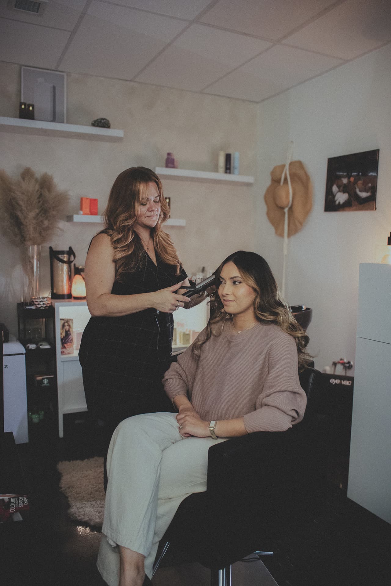 Steph styling a seated guest's long hair with a flat iron in The Sanctuary's warm, softly lit studio
