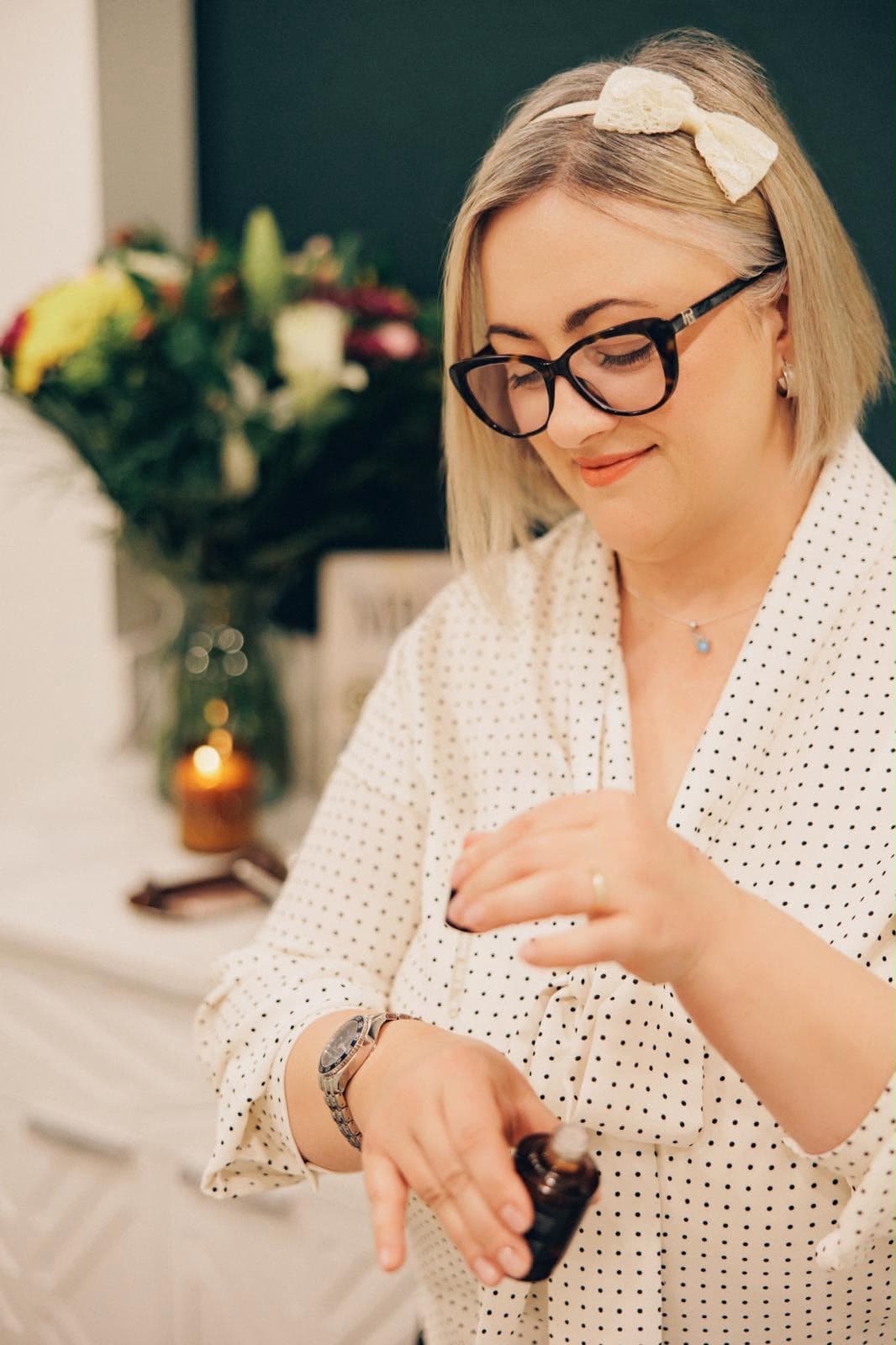 Mihaela of M Beauty and Artistry, in glasses and a polka-dot blouse, applying a drop of skincare oil to her wrist